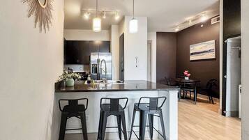 Una cocina moderna con una isla central, taburetes de bar, electrodomésticos de acero inoxidable, iluminación colgante y un comedor con mesa y sillas.