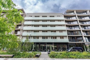 A multi-story building with a central entrance, surrounded by greenery and a paved area.