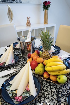 A dining table with a variety of fruits, a bottle of champagne, and a small figurine.
