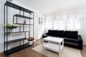 A living room with a black sofa, white coffee table, and a black metal shelving unit with flowers.