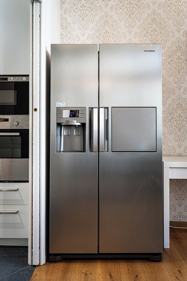 Un refrigerador de acero inoxidable con dispensador de agua y compartimento congelador, situado en una cocina con papel tapiz estampado y piso de madera.