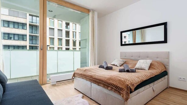Un dormitorio moderno con una cama grande, un ventanal de vidrio que da a un balcón y vista a un edificio de enfrente.