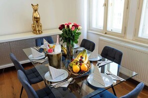 Un coin repas avec une table en verre, des chaises bleues, une statuette de chat dorée, un vase de fleurs et un panier de fruits.