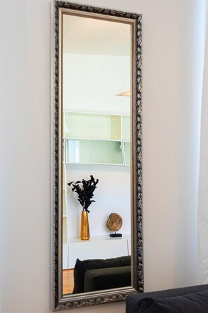 A tall, ornate mirror with a decorative frame, reflecting a room with a white wall, a black sofa, and a shelf with decorative items.