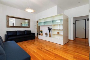 Un salon moderne avec un canapé noir, un plancher en bois, un meuble blanc avec des étagères en verre et un miroir au mur.