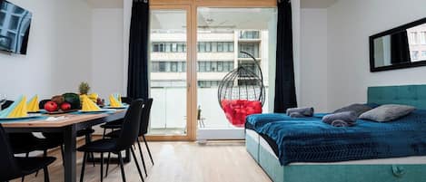 A modern bedroom with a bed, a dining table with chairs, a hanging chair, and a view of a building through a glass door.