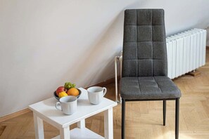 Une chaise grise, recouverte de tissu, se trouve à côté d’une table blanche sur laquelle est posé un saladier rempli de fruits et deux tasses blanches.