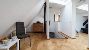 A modern, open-plan living space with a herringbone-patterned wooden floor, a grey upholstered chair, a wooden dresser, and a skylight above a staircase. - Royal Skyline Residence (Prague 5)