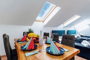 A dining area with a wooden table set for four, a fruit bowl, and a bottle of water. There are two blue upholstered chairs and a dark sofa in the background.