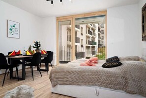 A modern bedroom with a large bed, a dining area with chairs, and a view of an apartment building through a glass door.