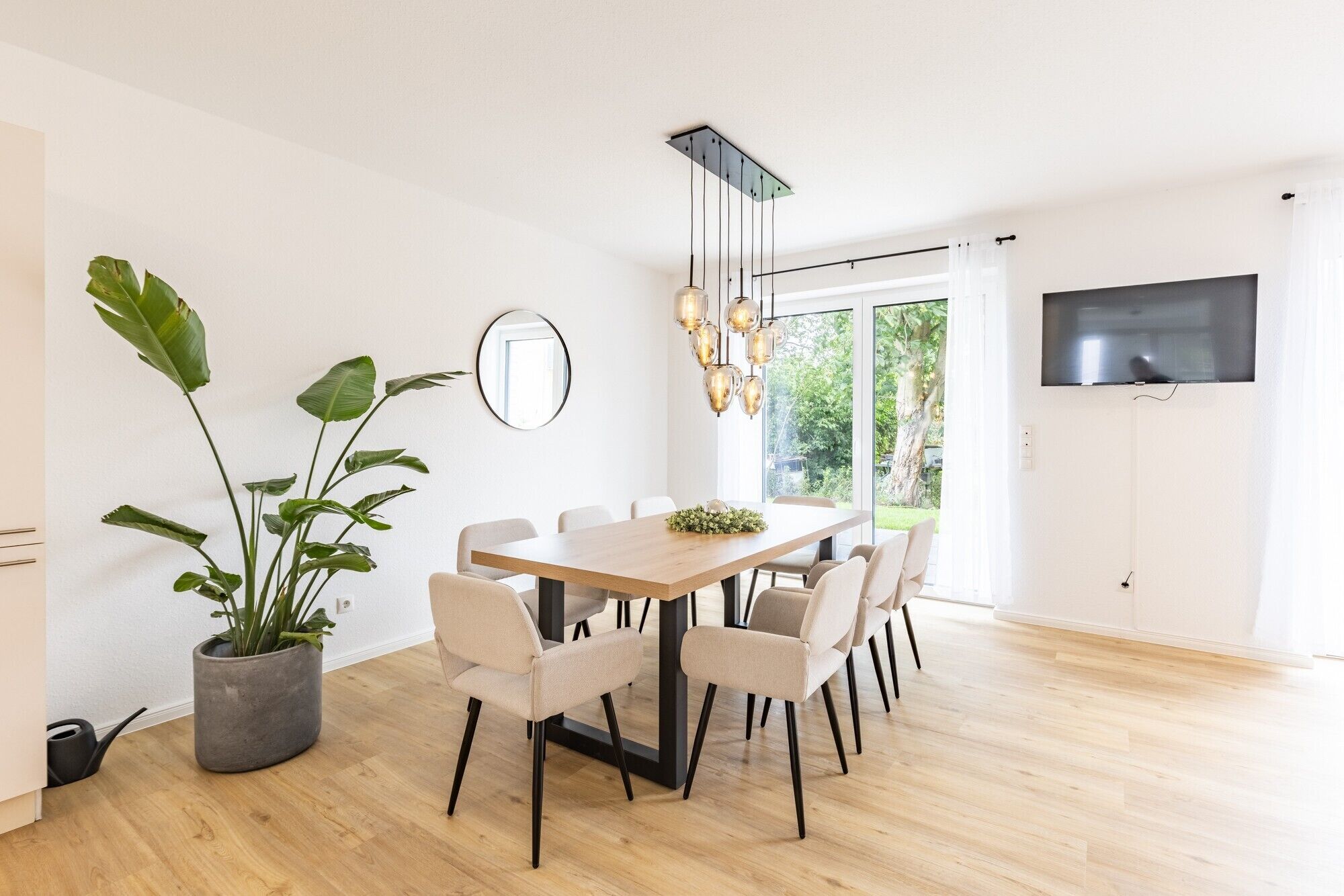 Un espace repas moderne avec une table en bois, des chaises blanches, une plante en pot et un grand miroir.