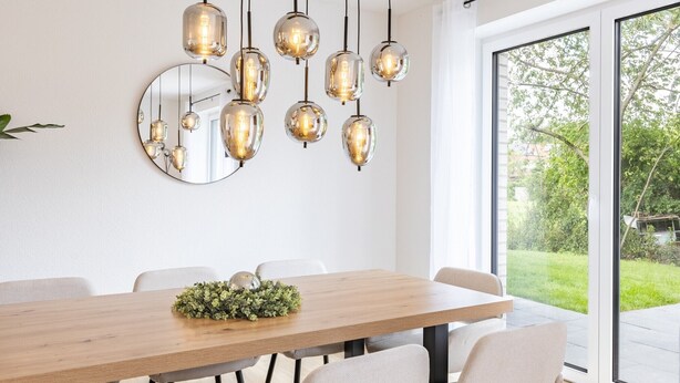 A dining area with a wooden table, multiple chairs, hanging pendant lights, a round mirror, and a view of a green outdoor area through a large window. - Holiday Home 'Das Strandhaus 2' with Shared Terrace and Wi-Fi (Lembruch)
