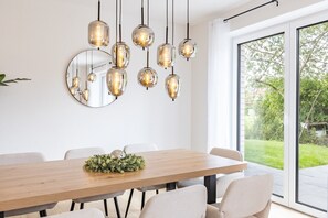 A dining area with a wooden table, multiple chairs, hanging pendant lights, a round mirror, and a view of a green outdoor area through a large window.