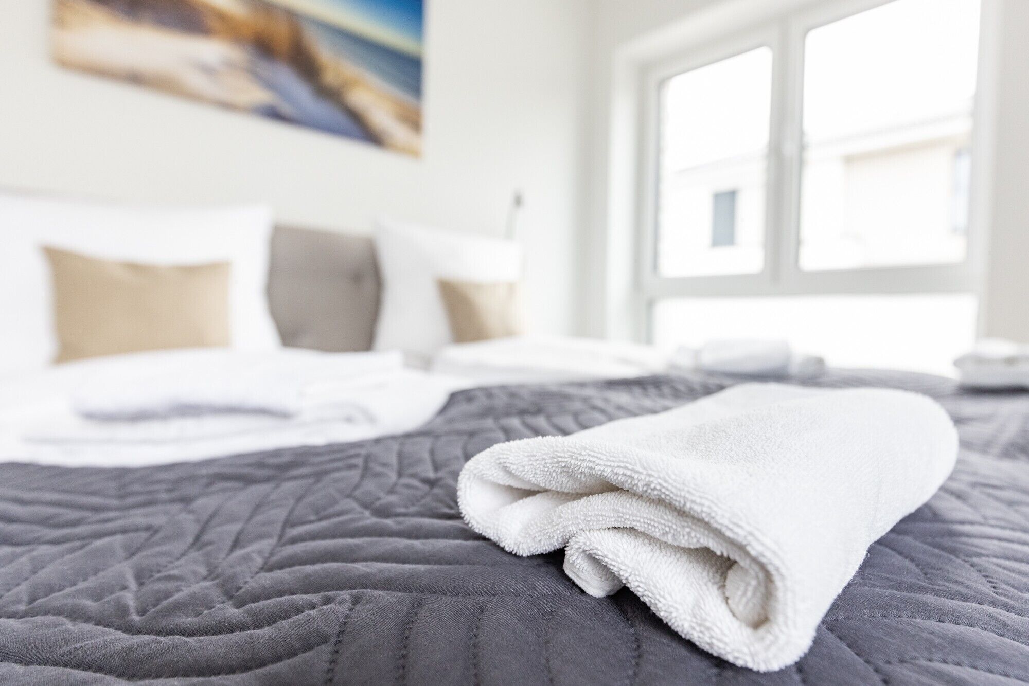 Una cama bien tendida con una toalla blanca doblada, una colcha gris acolchada y una ventana con luz del día.