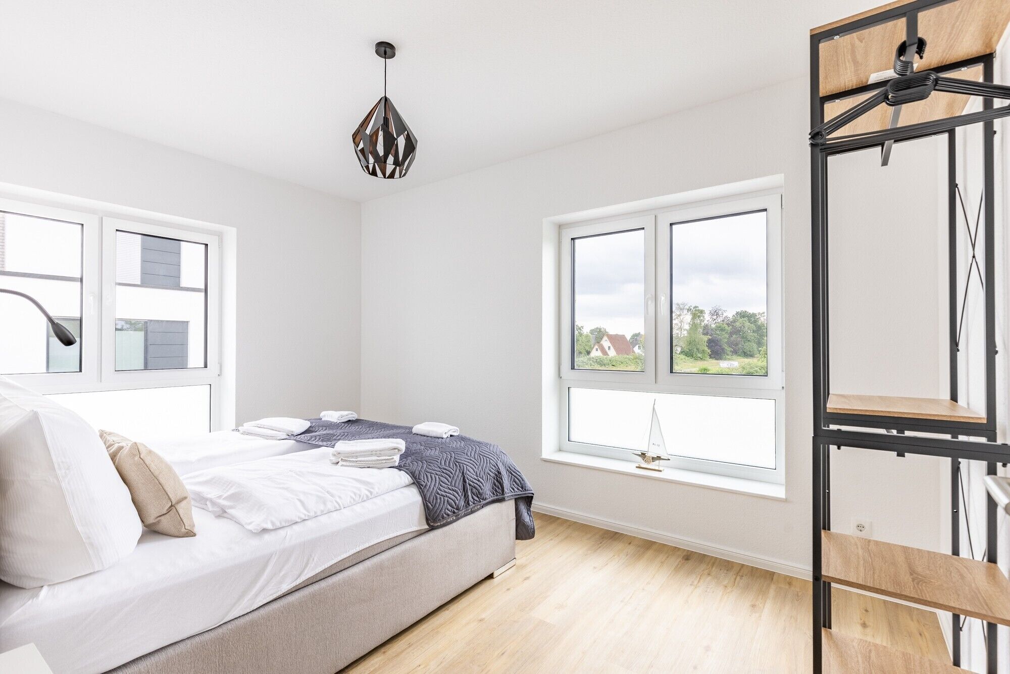 Een slaapkamer met een bed, twee ramen, een hanglamp en een houten wandplank.