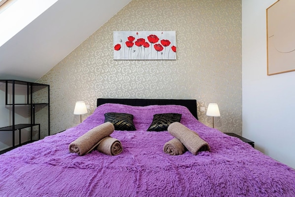 A bedroom with a large bed, two pillows, a wall painting, and a nightstand with a lamp.