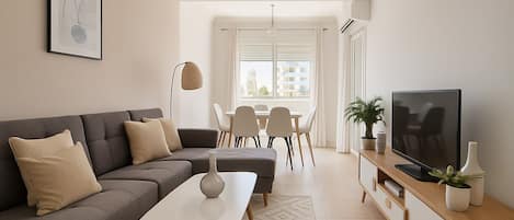 A modern living room with a grey sofa, a white coffee table, a TV on a wooden stand, and a dining area with chairs and a table.