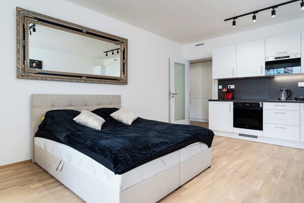 A modern bedroom with a large bed, a mirror, and a kitchenette with a stove and coffee maker.