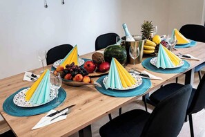 Uma mesa de jantar posta com jogos americanos azuis, uma fruteira, uma garrafa de champanhe e uma melancia.
