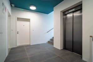 Un couloir avec une porte blanche, un escalier et un ascenseur métallique.