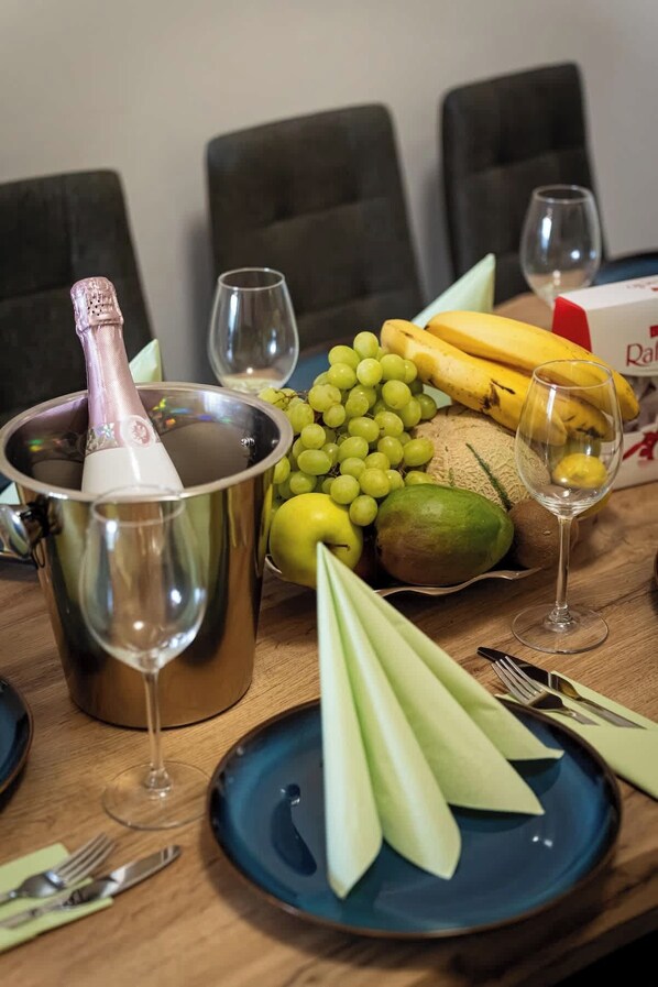 A dining table set with a champagne bottle, wine glasses, a bowl of grapes, bananas, apples, and a plate with cut vegetables.