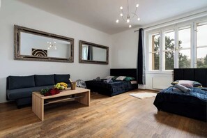 A modern room with a dark sofa, a bed, and a wooden coffee table with fruits and flowers. There are two large mirrors on the wall and a window with curtains. - Royal Aurora Residence (Prague 7)