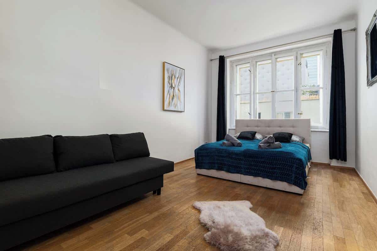 Une chambre à coucher avec un canapé noir, un lit recouvert d’une couverture bleue, une tête de lit blanche et une grande fenêtre ornée de rideaux noirs.