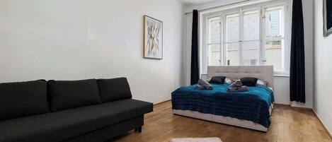 Une chambre à coucher avec un canapé noir, un lit recouvert d’une couverture bleue, une tête de lit blanche et une grande fenêtre ornée de rideaux noirs.