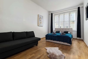 A bedroom with a black sofa, a bed with a blue blanket, a white headboard, and a large window with black curtains.