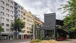 Un edificio moderno con fachada de vidrio, una pequeña zona de estar al aire libre y una calle frente a él.
