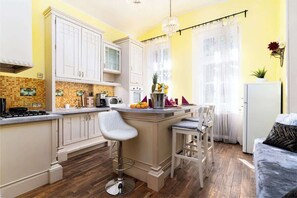 A kitchen with white cabinets, a central island with bar stools, a refrigerator, and a window with sheer curtains. - Imperial Deluxe Suite (Prague)