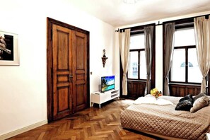 A bedroom with a wooden wardrobe, a television on a white stand, a bed with a quilted cover, and a window with curtains. - Imperial Deluxe Suite (Prague)