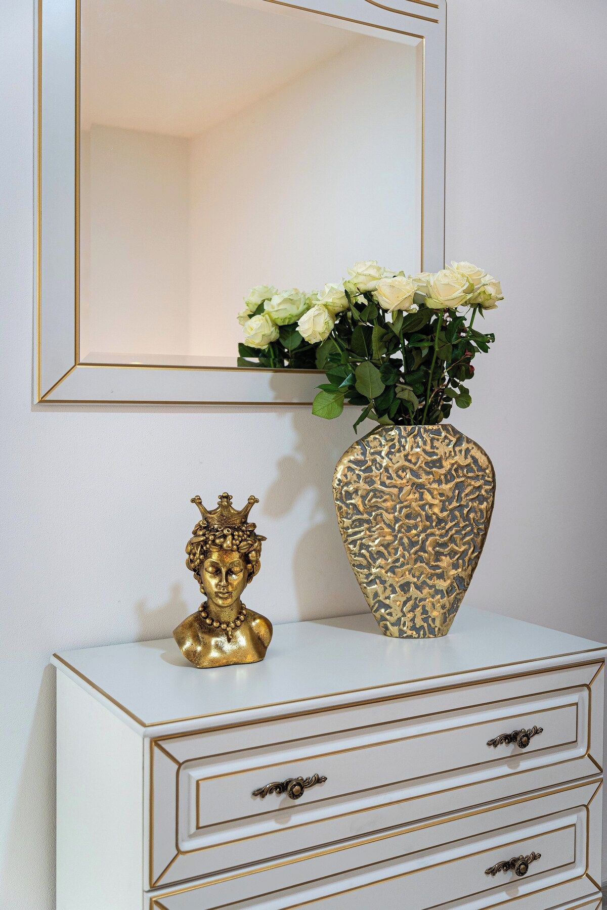 Ein weißer Schminktisch mit goldener Zierleiste und Schubladen, eine goldene Büste und eine Vase mit weißen Blumenmuster.