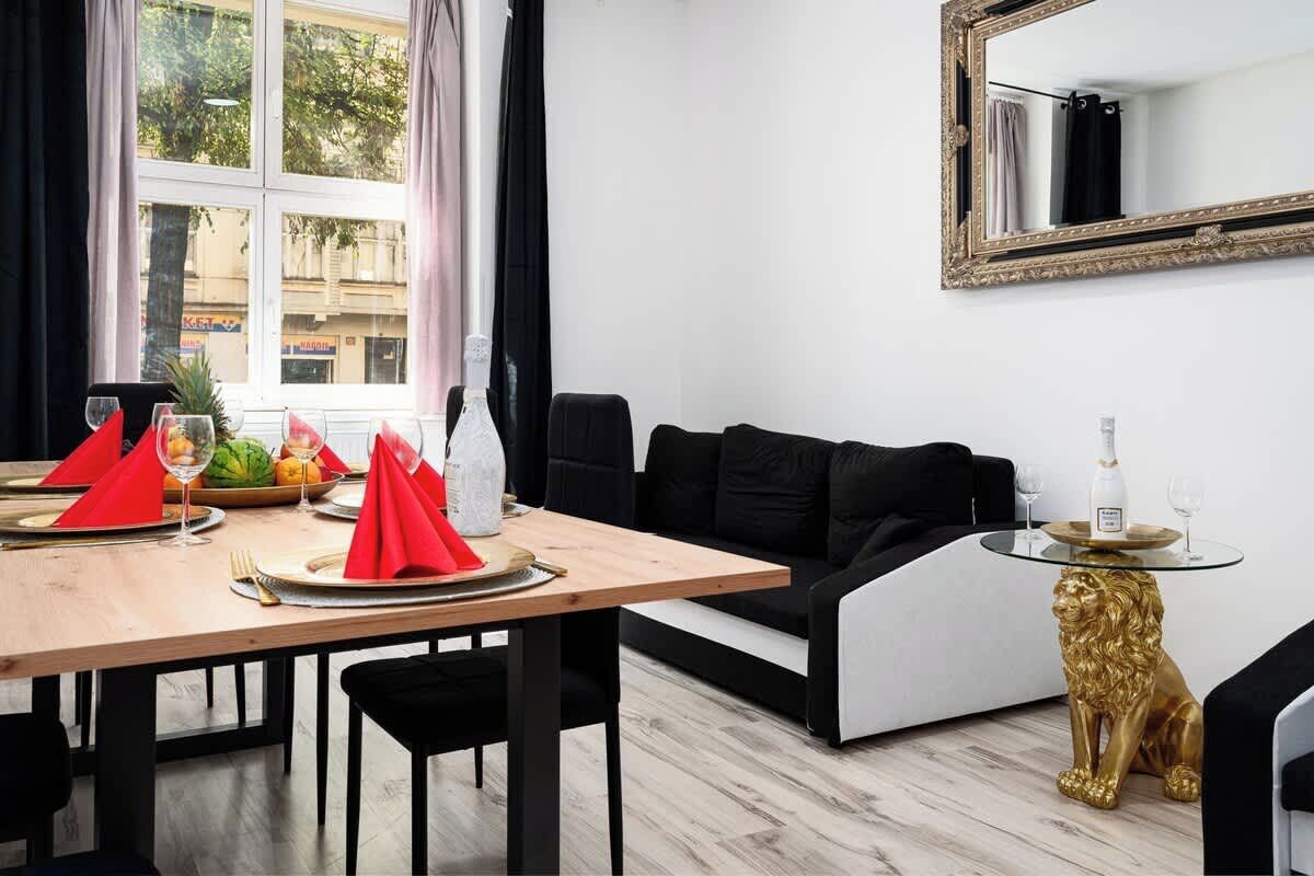 Un coin repas avec une table en bois mise pour deux personnes, des serviettes rouges et un verre d’eau. Un canapé noir et une statue de lion dorée se trouvent à côté de la table.