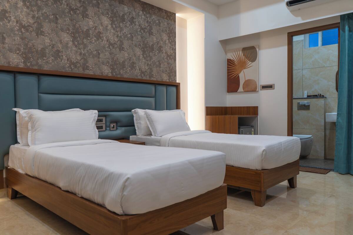 A hotel room with two beds, a wooden headboard, a small bathroom with a shower, and a large window with curtains.