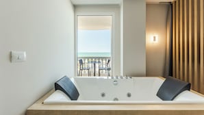 A modern bathroom with a large bathtub, a window with a view of the sea, and a wooden panel wall. - SEA LA VIE SUITES (Golem)