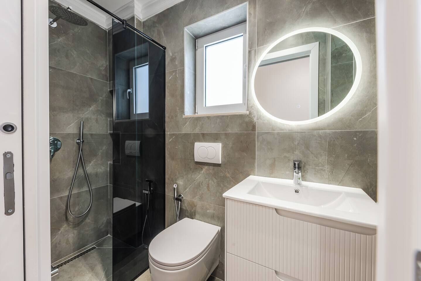 A modern bathroom with a glass-enclosed shower, a toilet, a sink with a mirror, and a window providing natural light.