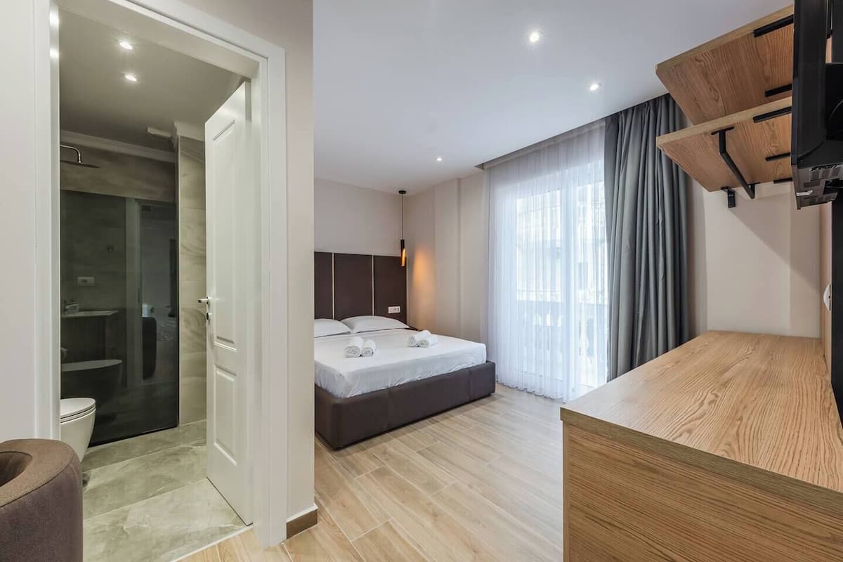 Una habitación de hotel moderna con una cama, un escritorio de madera, zona de ducha y un amplio ventanal con cortinas.