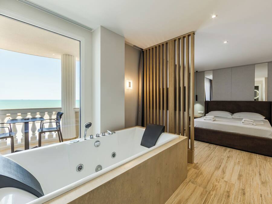 A modern bedroom with a large bathtub, a bed with neatly made linens, and a view of the ocean through a balcony.