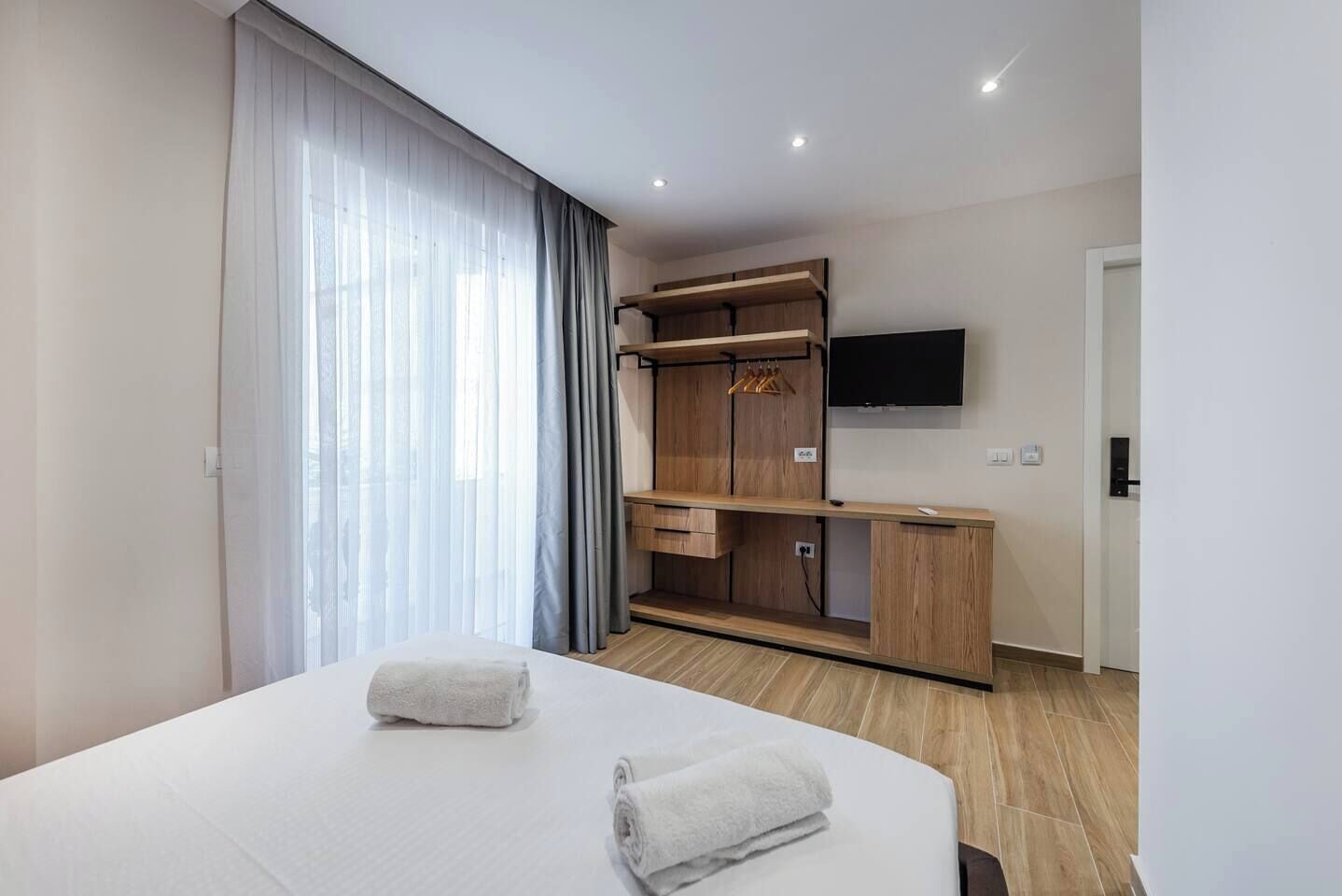 Quarto de hotel moderno com cama grande, guarda-roupa de madeira, televisão de tela plana e janela com cortinas translúcidas.