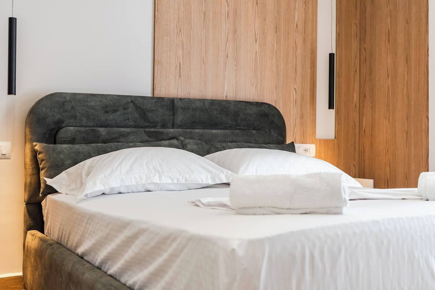 Een net opgemaakt bed met witte lakens en kussens, een donkergrijze beklede hoofdbord en houten paneelbekleding aan de muur.
