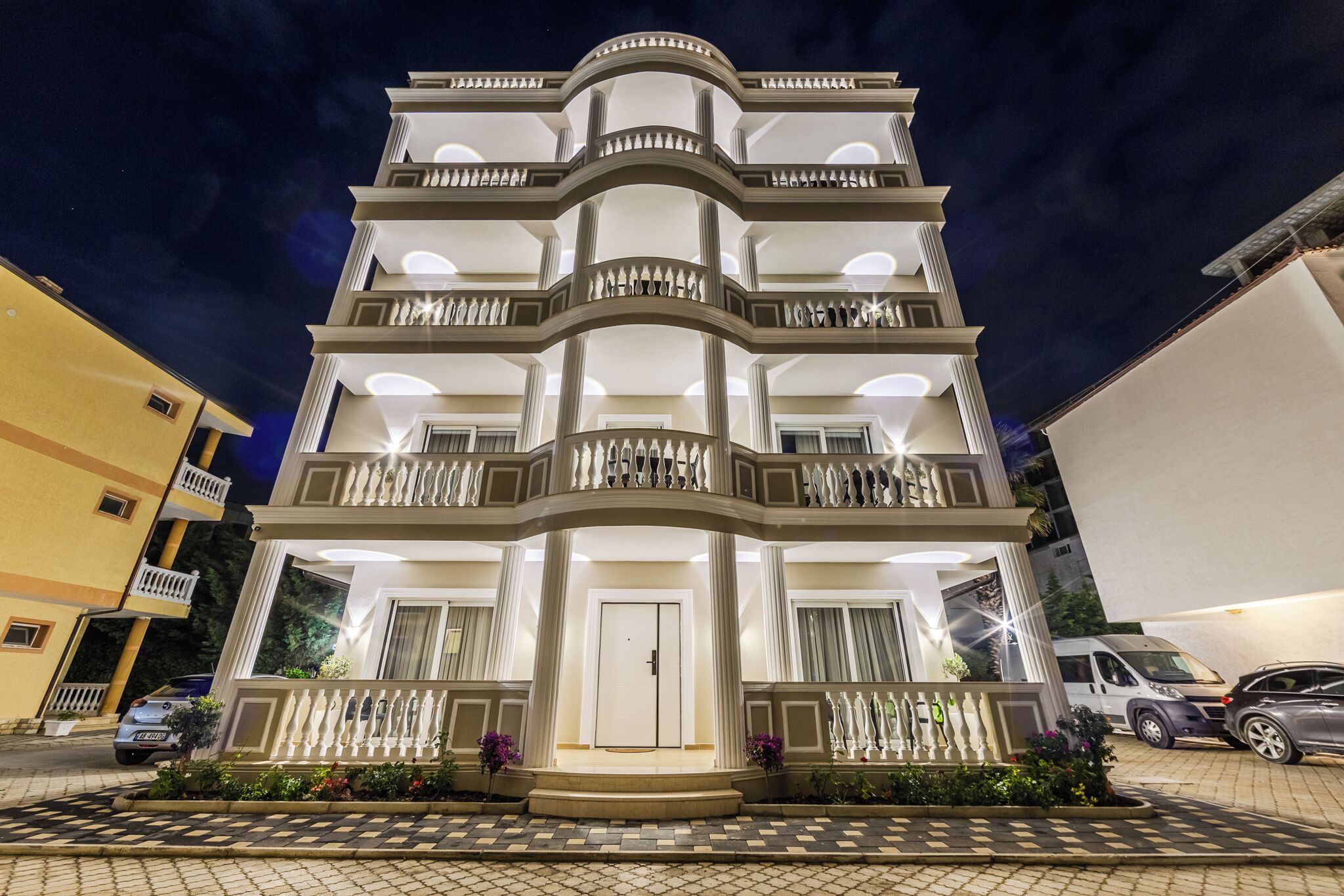 A multi-story building with balconies, a central entrance, and a parking area.