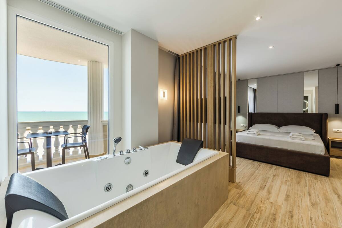 Un dormitorio moderno con una bañera grande, una cama con ropa de cama impecable y vistas al océano desde un balcón.