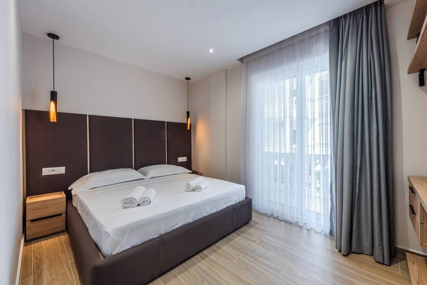 Una habitación de hotel moderna con una cama grande, cabecera de madera, mesitas de noche y vistas de la ciudad a través de cortinas traslúcidas.