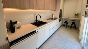 A modern kitchen with white cabinets, a black sink, and a built-in oven. There are two stools and a vase with flowers on the counter.