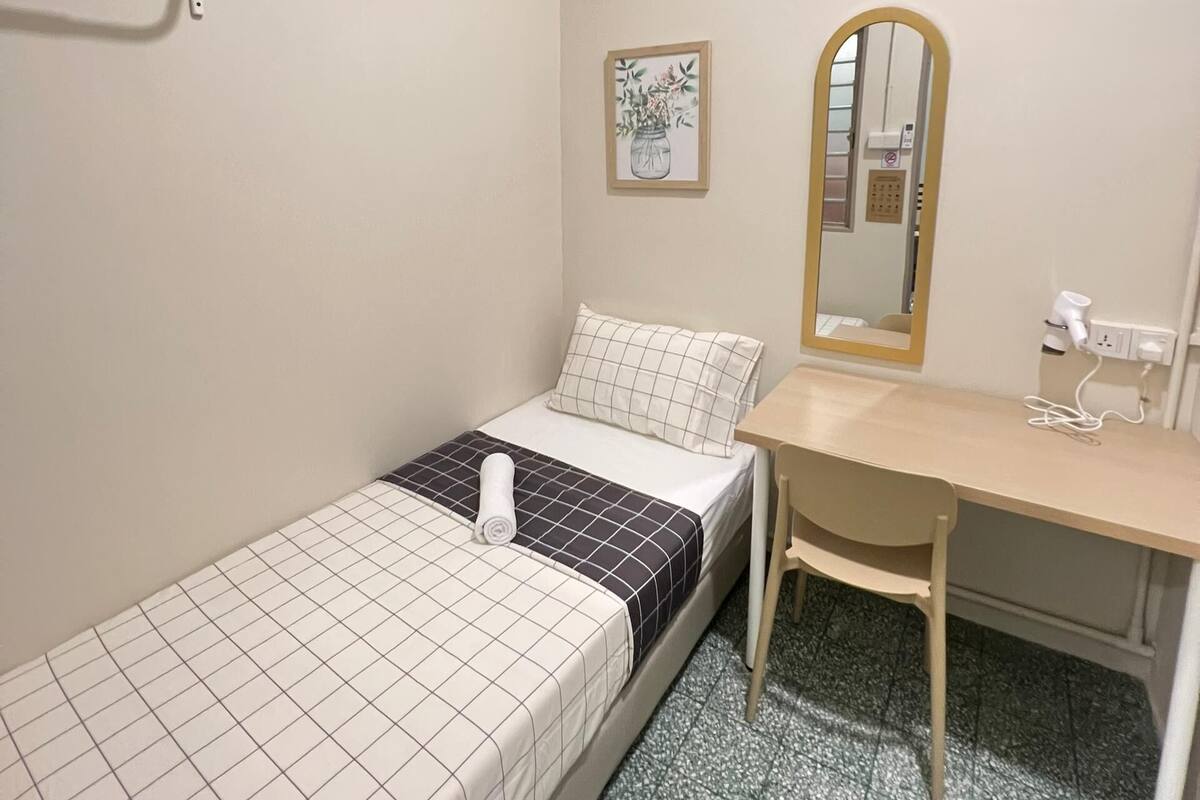 A small, single-bed room with a desk, chair, and an air conditioning unit.