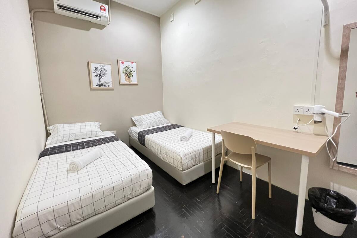 A small, clean room with two beds, a desk, and an air conditioning unit.