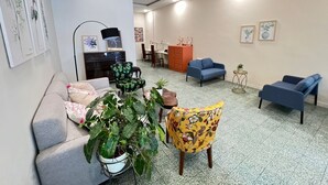 A spacious living area with a grey sofa, a yellow armchair, and a blue sofa. There are several potted plants and a small table with a plant. The room has a tiled floor and white walls with framed artwork. - myRoom at 14 J Mandalay (George Town)