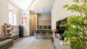 Una cocina moderna con una isla central, un comedor con taburetes de bar y una sala de estar con sofá y estantería.