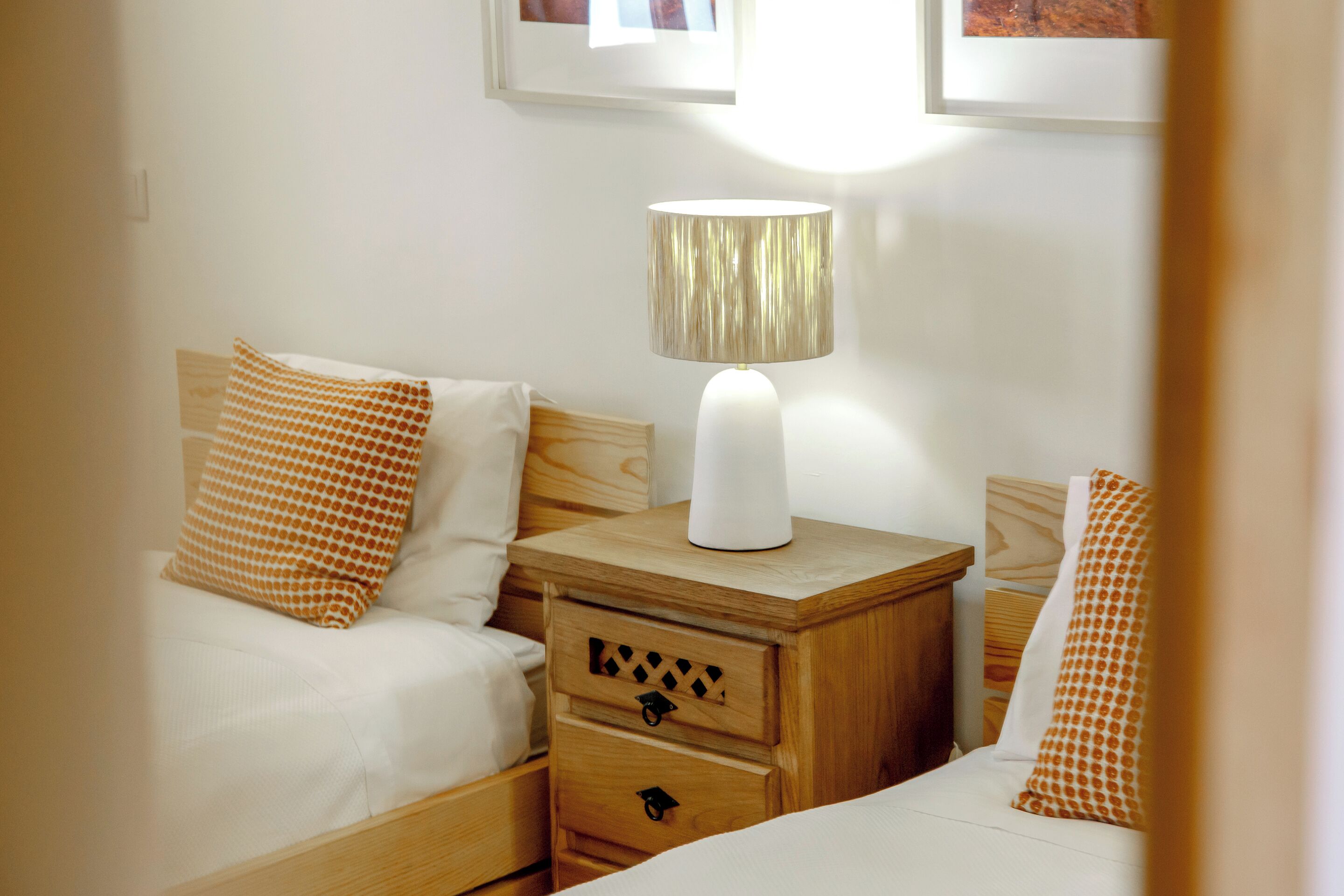 Una cama doble con ropa blanca y almohadas con estampado naranja, una mesita de noche de madera con una lámpara y un cajón.